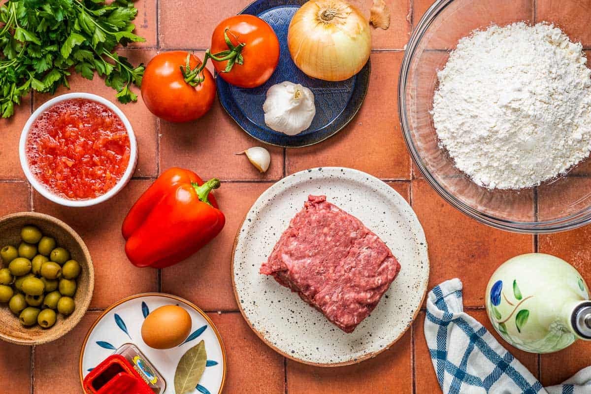 ingredients for spanish beef empanadillas including ground been, olive oil, flour, onion, tomatoes, garlic, red pepper, grated tomatoes, an egg, bay leaf, sweet paprika, and green olives.
