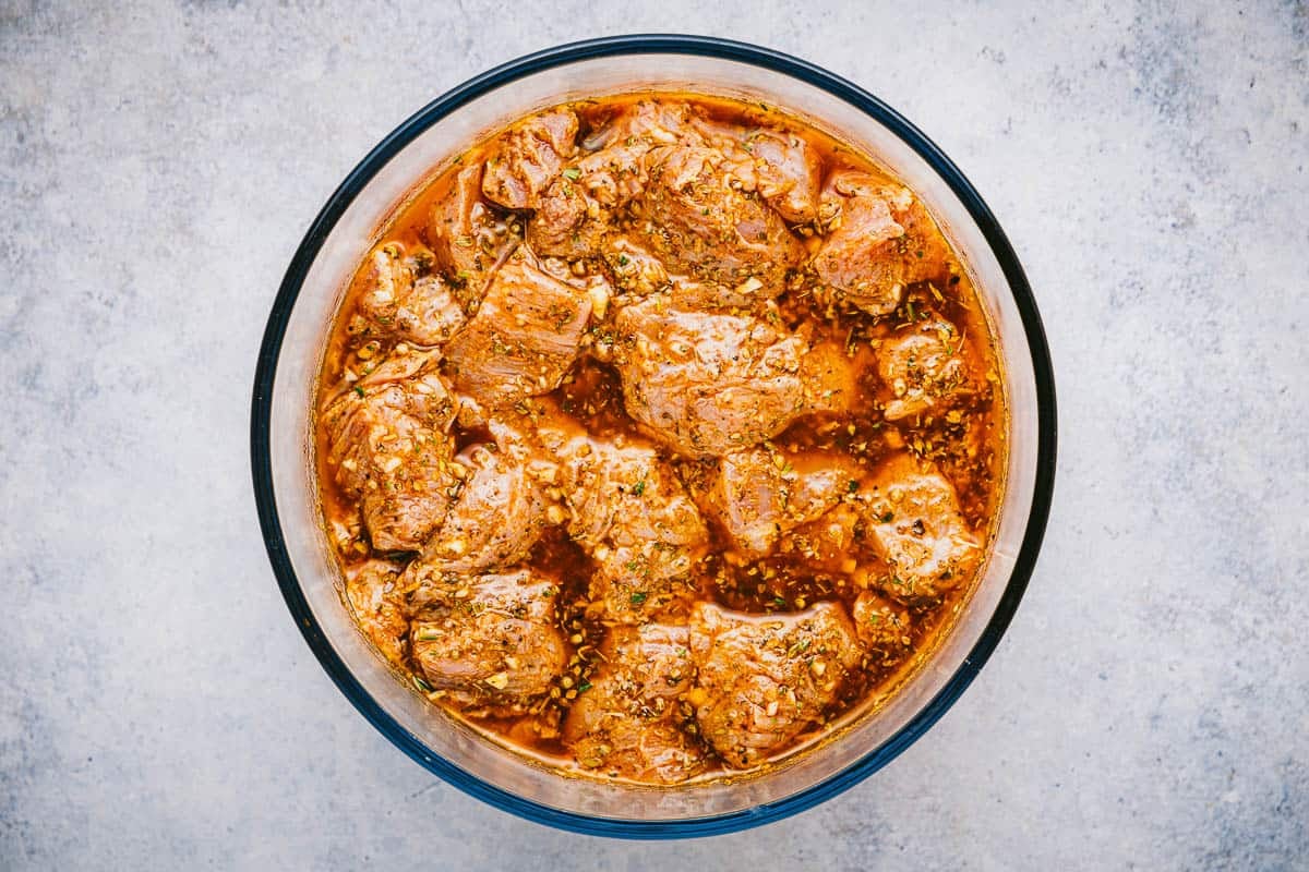 Pork marinating in a bowl with white wine vinegar, olive oil, garlic cloves, mustard, oregano, rosemary, paprika, and black pepper to make pork souvlaki.