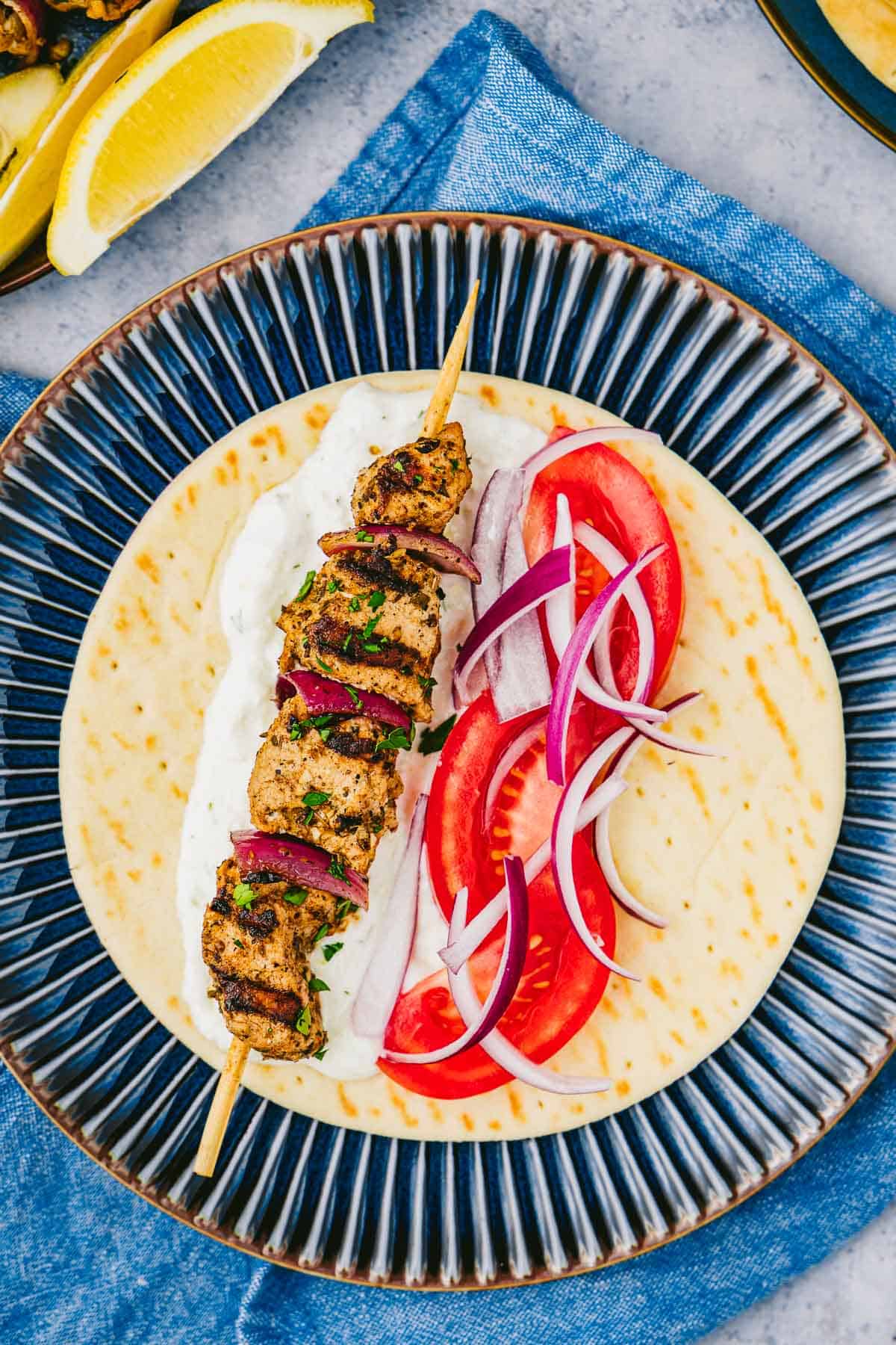 Overhead shot of a pita bread with tzatziki, red onion, tomato, and a pork souvlaki skewer.