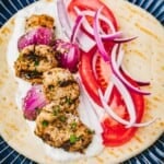 Side shot of a pita bread with tzatziki, red onion, tomato, and pork souvlaki on top.