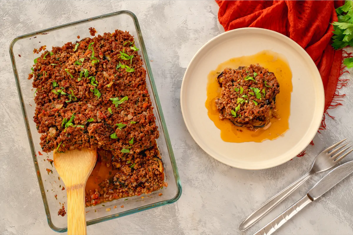 Freshly baked Moussaka served on a plate from a baking dish containing more of the recipe.