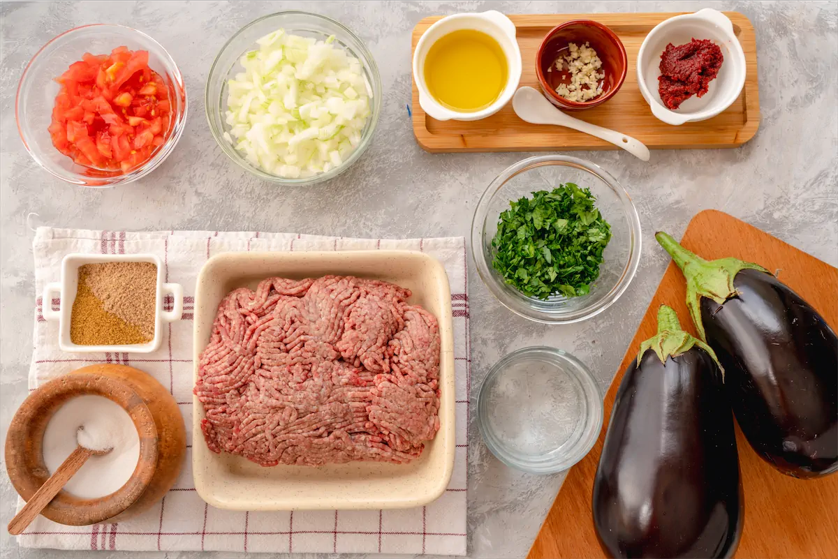 Large bell eggplants, ground beef, olive oil, chopped onion, minced garlic, water, tomatoes, tomato paste, salt, black pepper, cayenne, cumin, and chopped parsley made ready on the kitchen table.