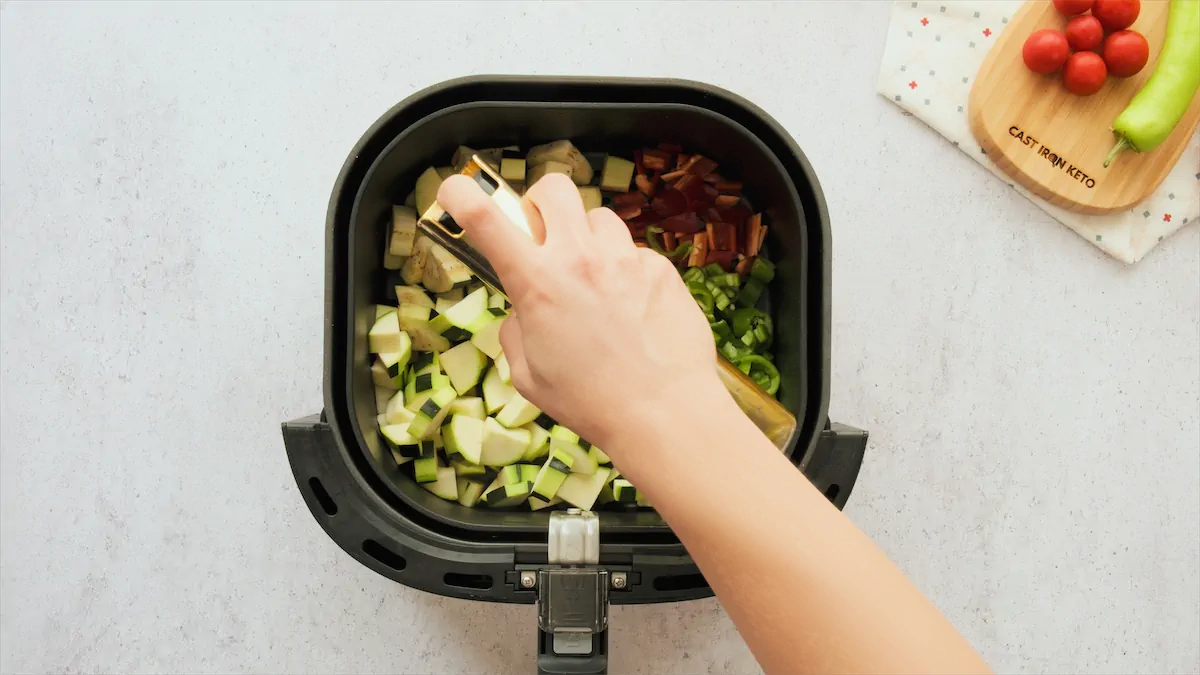 Drizzling olive oil over chopped eggplant, zucchini, and bell pepper in an air fryer basket.