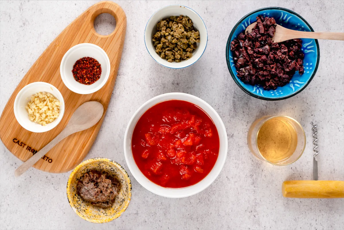 Diced tomatoes, chopped anchovy fillets, minced garlic cloves, chopped capers, pitted and chopped black olives, cup of dry white wine, and chilli flakes made ready on the kitchen table.