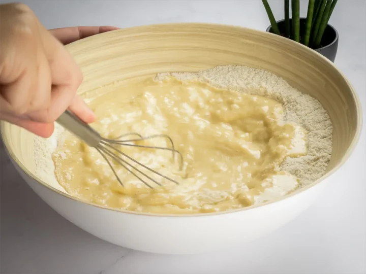 A hand mixing wet and dry ingredients with a whisk in a bowl.