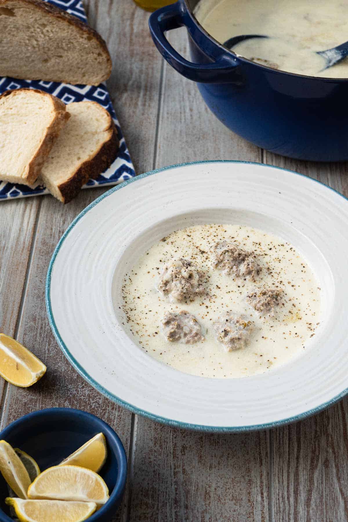 Youvarlakia Greek Meatball Soup in a bowl surrounded by a small bowl of lemon wedges, a plate of sliced bread and a pot of the soup.