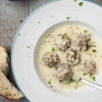 Overhead photo of Youvarlakia Greek Meatball Soup in a bowl surrounded by a small bowl of lemon wedges, bowls of salt and pepper, and a piece of crusty bread.