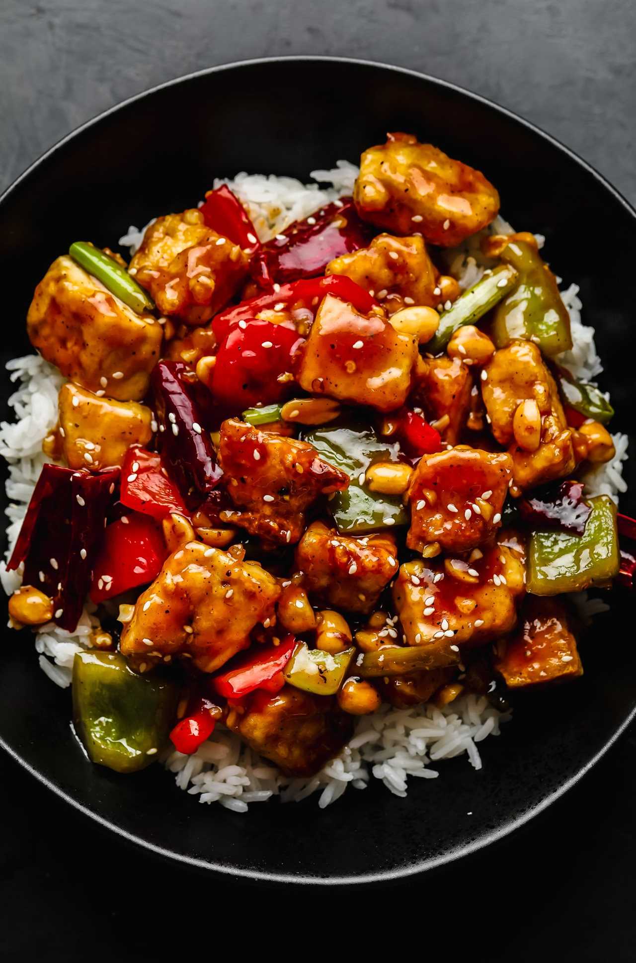 vegan kung pao tofu and vegetables served on a bed of rice in a black bowl.