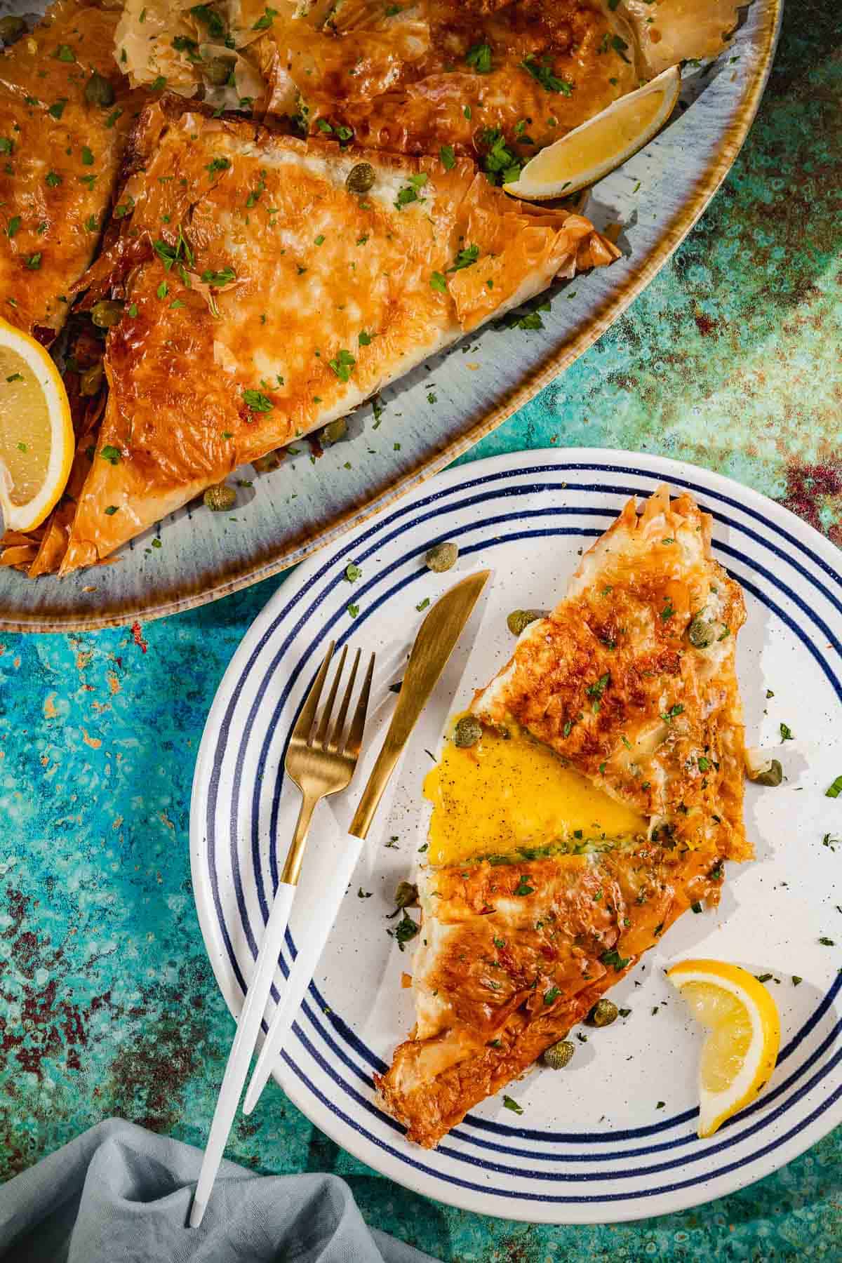 one piece of brik cut in half on a plate with a knife, for, and a lemon wedge in front of a platter containing several pieces of brik.