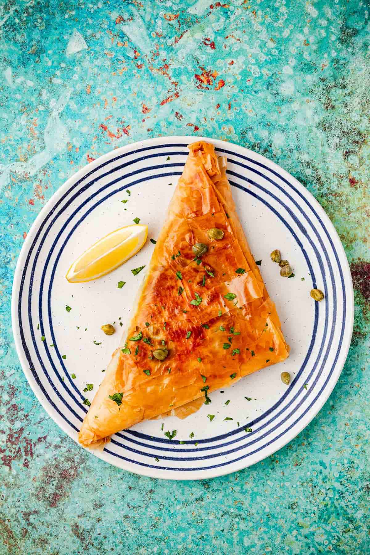 an overhead photo of a cooked piece of brik garnished with chopped parsley and capers on a plate with a lemon wedge.