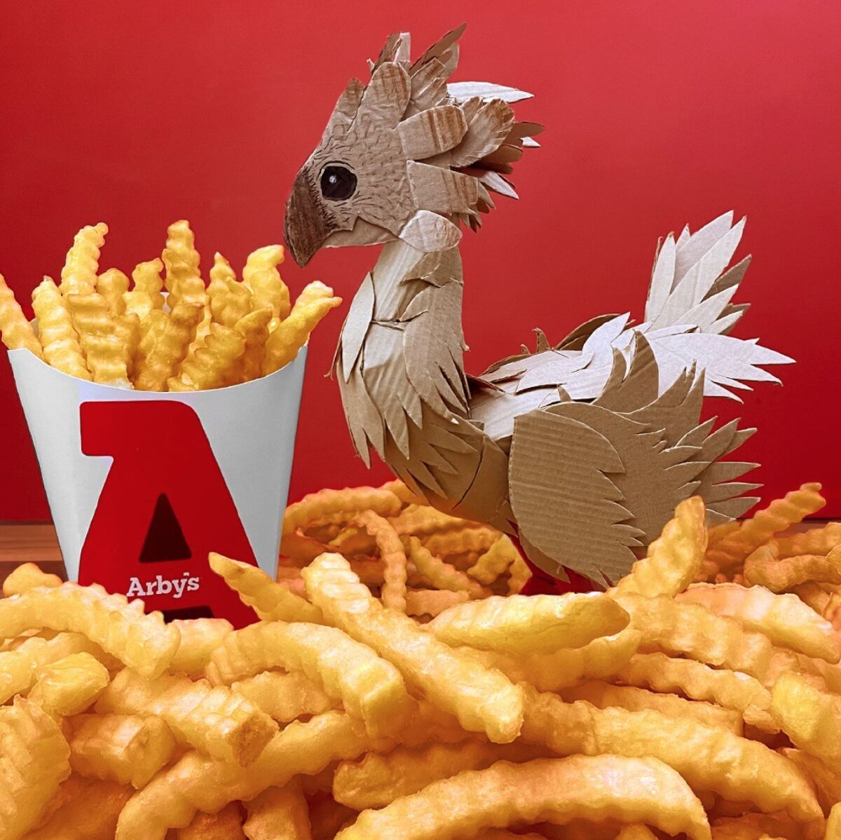 A white and red box of Arby's crinkle fries next to a brown and white feathered bird amongst a table full of fries on a red background. 