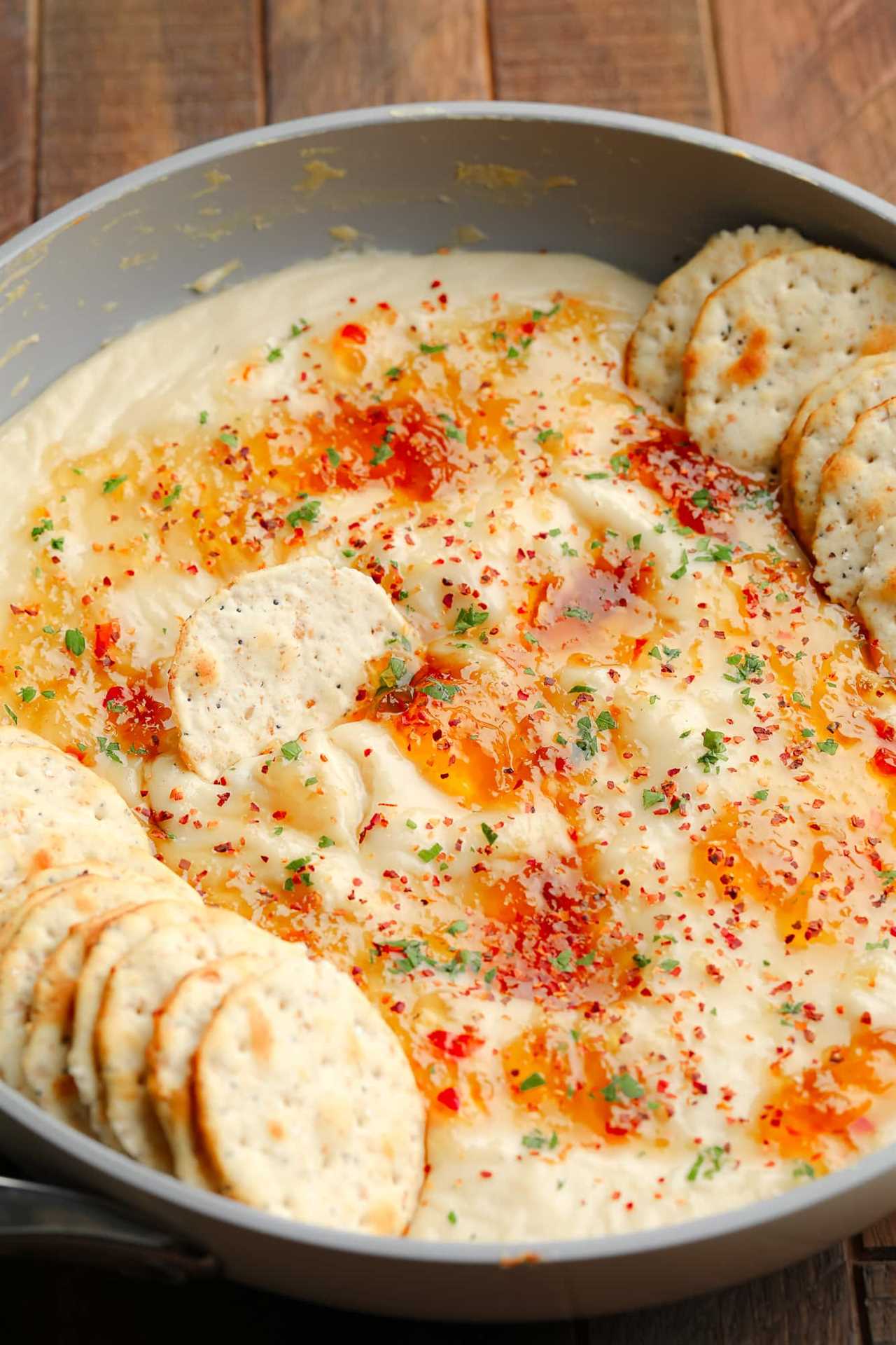 baked vegan brie dip topped with pepper jelly and crackers in a grey saucepan.