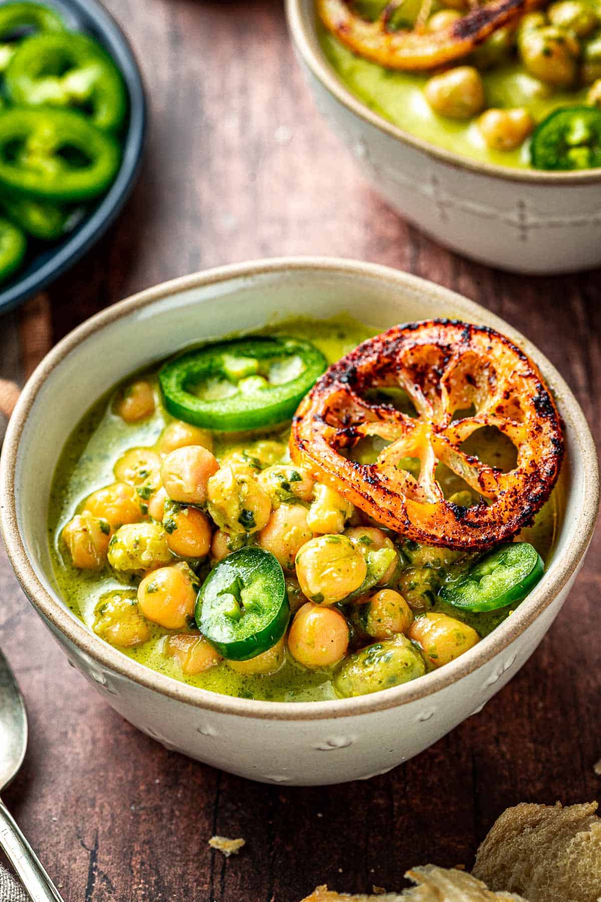 close up of two bowls of creamy pesto braised chickpeas topped with jalapeno and fried lemon slices, next to a plate of sliced jalapeno.