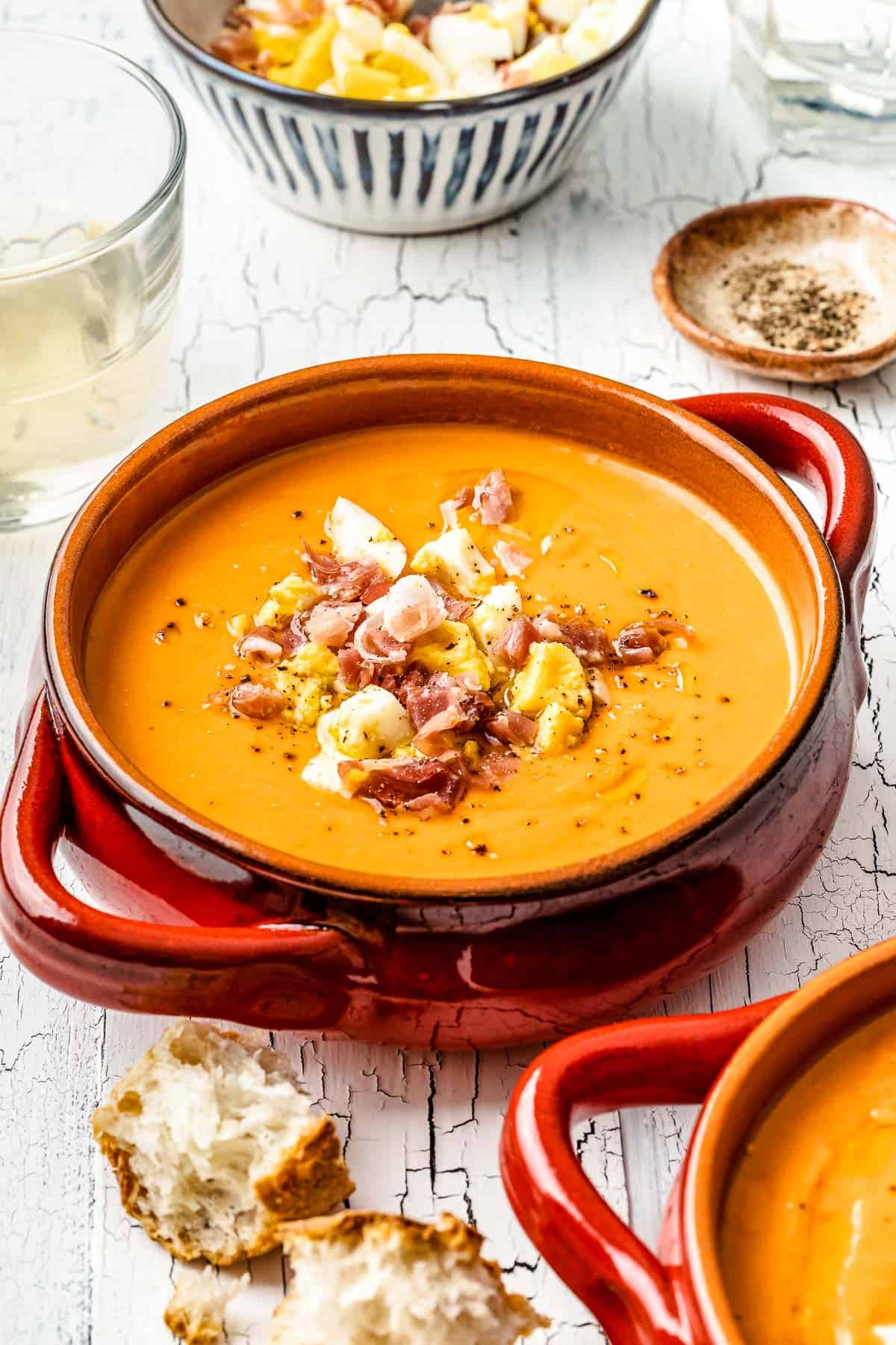 close up of a bowl of salmorejo spanish cold tomato soup topped with chopped hard boiled eggs and prosciutto next to a glass of wine and a bowl of pepper.