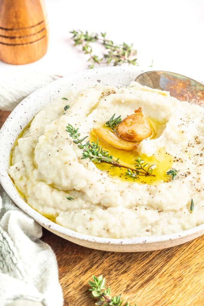 Keto Mashed Cauliflower in a white bowl topped with whole roasted garlic cloves, a sprig of Thyme, and some props to the side