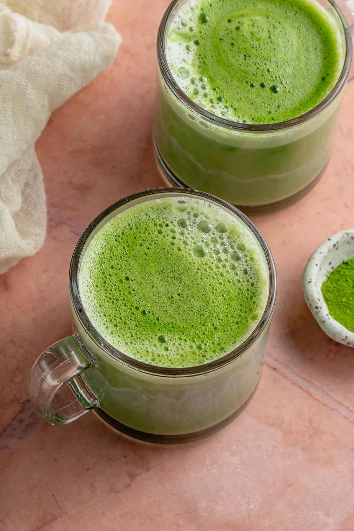 2 frothy matcha lattes in glass cups on pink table with white towel