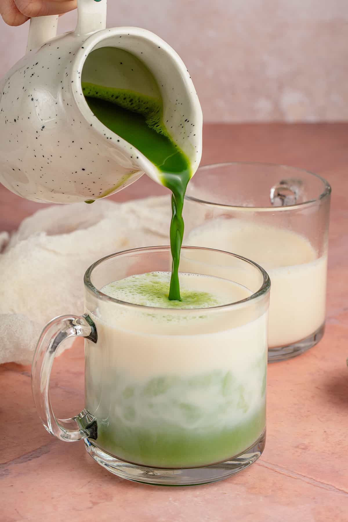 pouring whisked matcha into hot soy milk in glass cup for latte on pink table