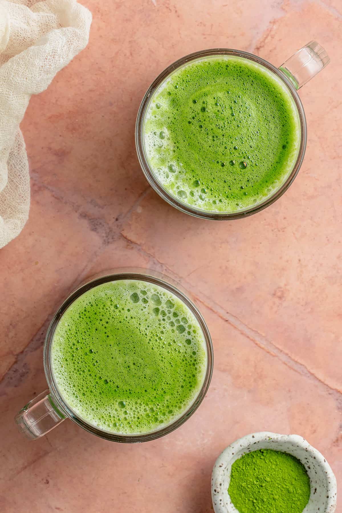 overhead photo of 2 frothy matcha lattes in glass cups on pink table with white towel