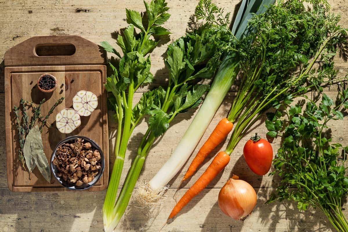 Ingredients for homemade vegetable stock including celery, carrots, a leek, onion, tomato, garlic, mushrooms, parsley, thyme, bay leaves, and black peppercorns.