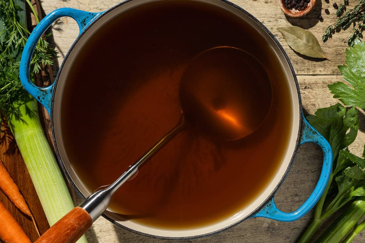 Homemade vegetable stock in a pot with a ladle. This is surrounded by carrots, a lee, celery, a bay leaf, thyme and a small bowl of black peppercorns.