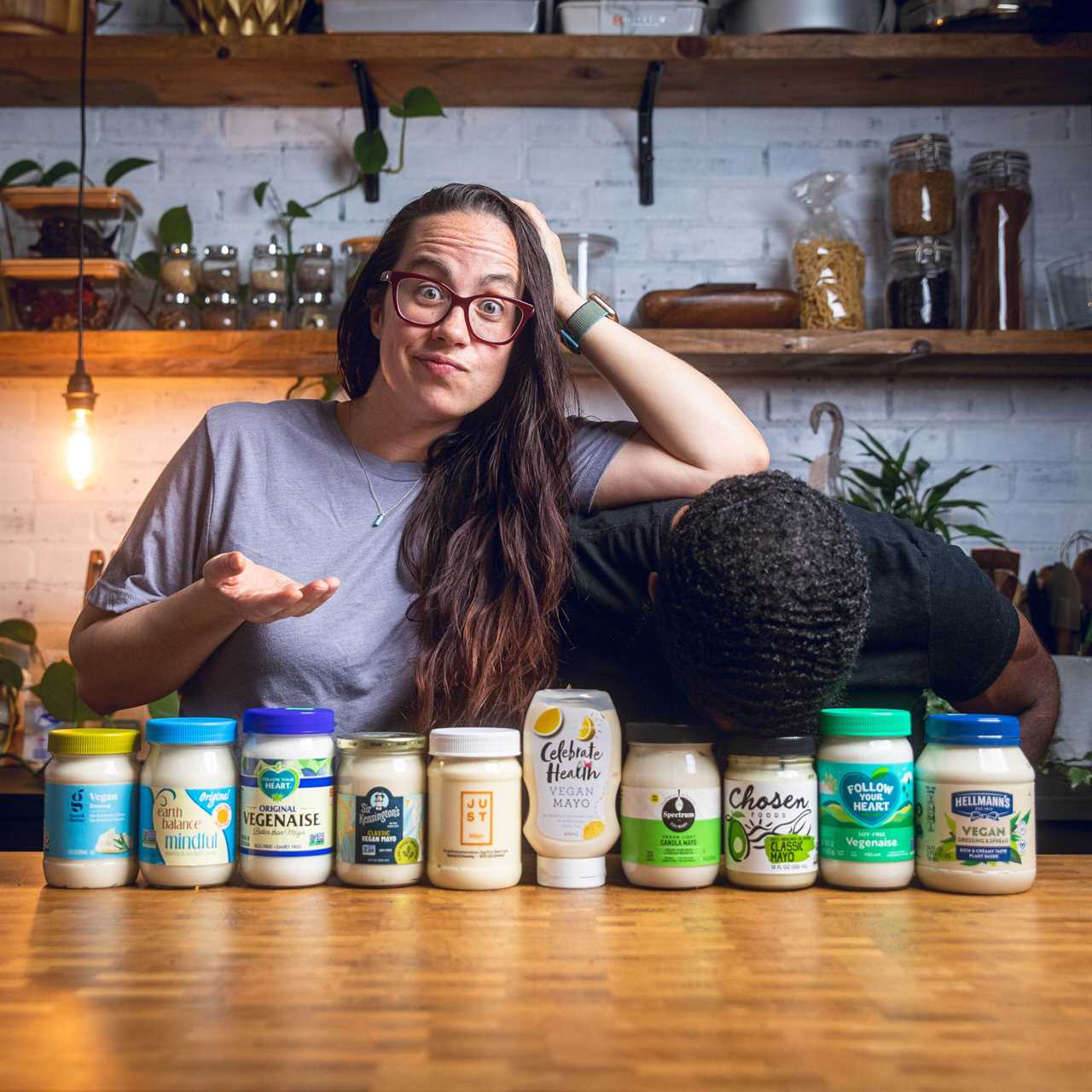 A couple posing funnily in front of an assortment of vegan mayo bottles.