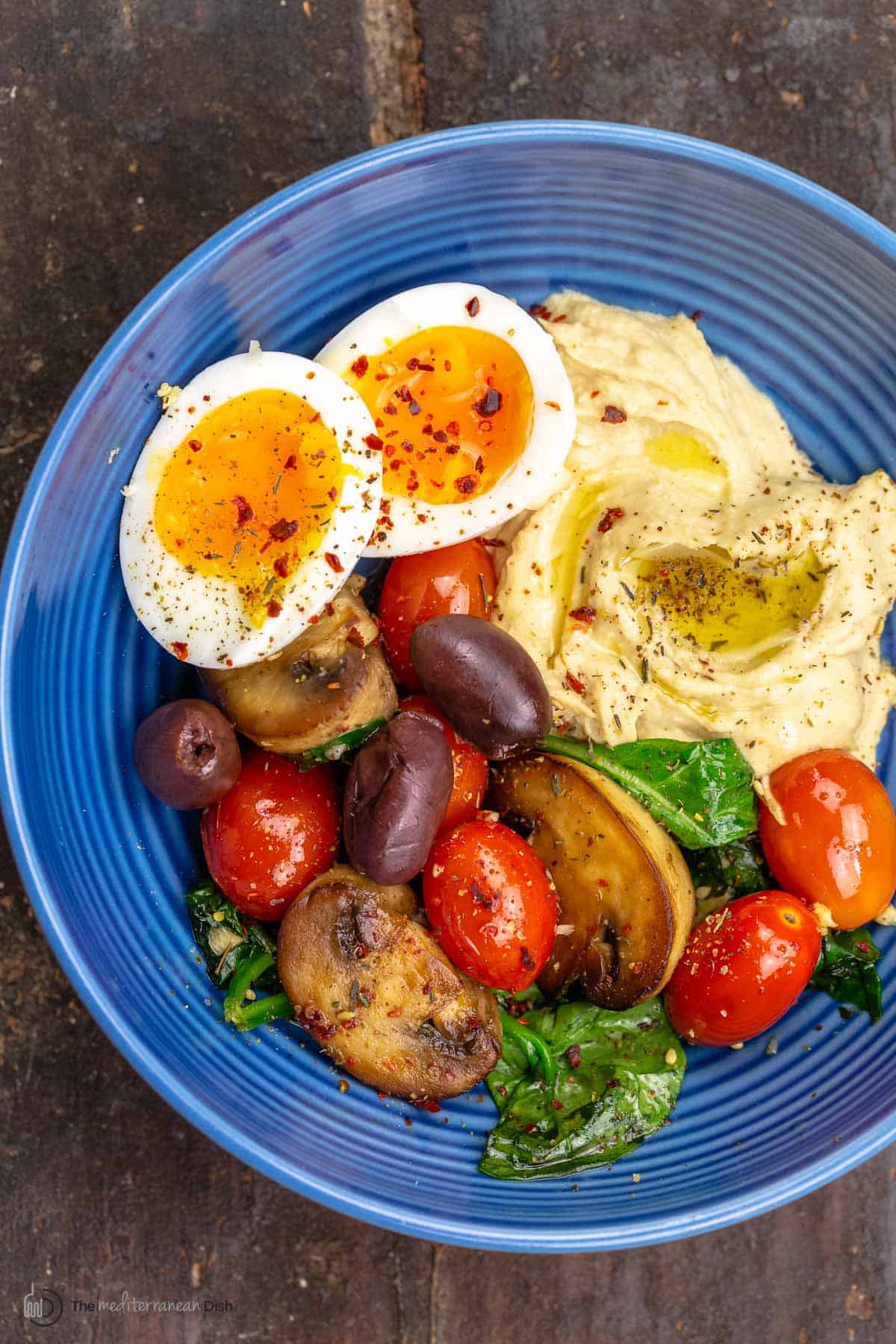 A Mediterranean breakfast bowl with a soft-boiled egg, hummus and sauteed vegetables on a blue plate.