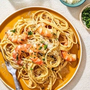 An overhead photo of a plate of cacio e pepe with a fork. Next to this is a glass of water and small bowls of grated pecorino romano cheese and parsley.