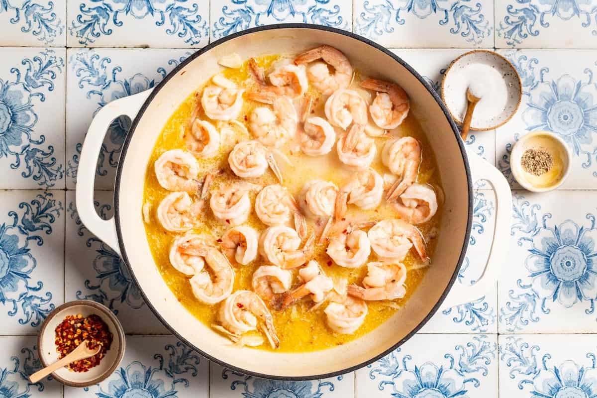 Shrimp cooking in the sauce in a large pot. Next to this are small bowls of salt, pepper and red pepper flakes.