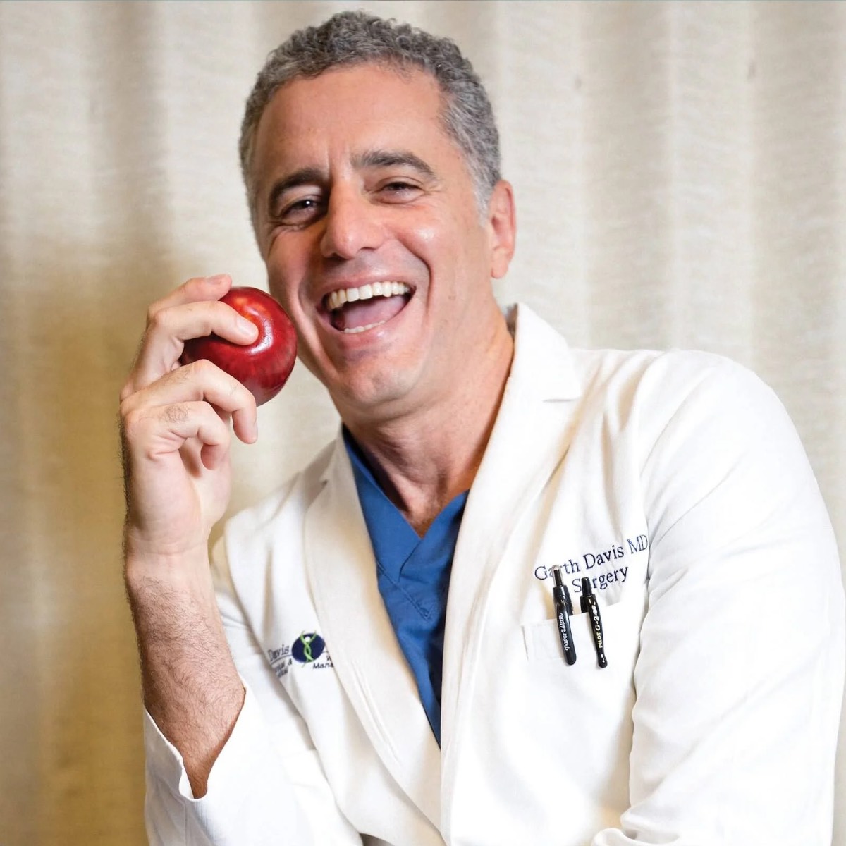 Headshot photo of plant-based vegan Dr. Garth Davis holding an apple in his doctor's coat. 
