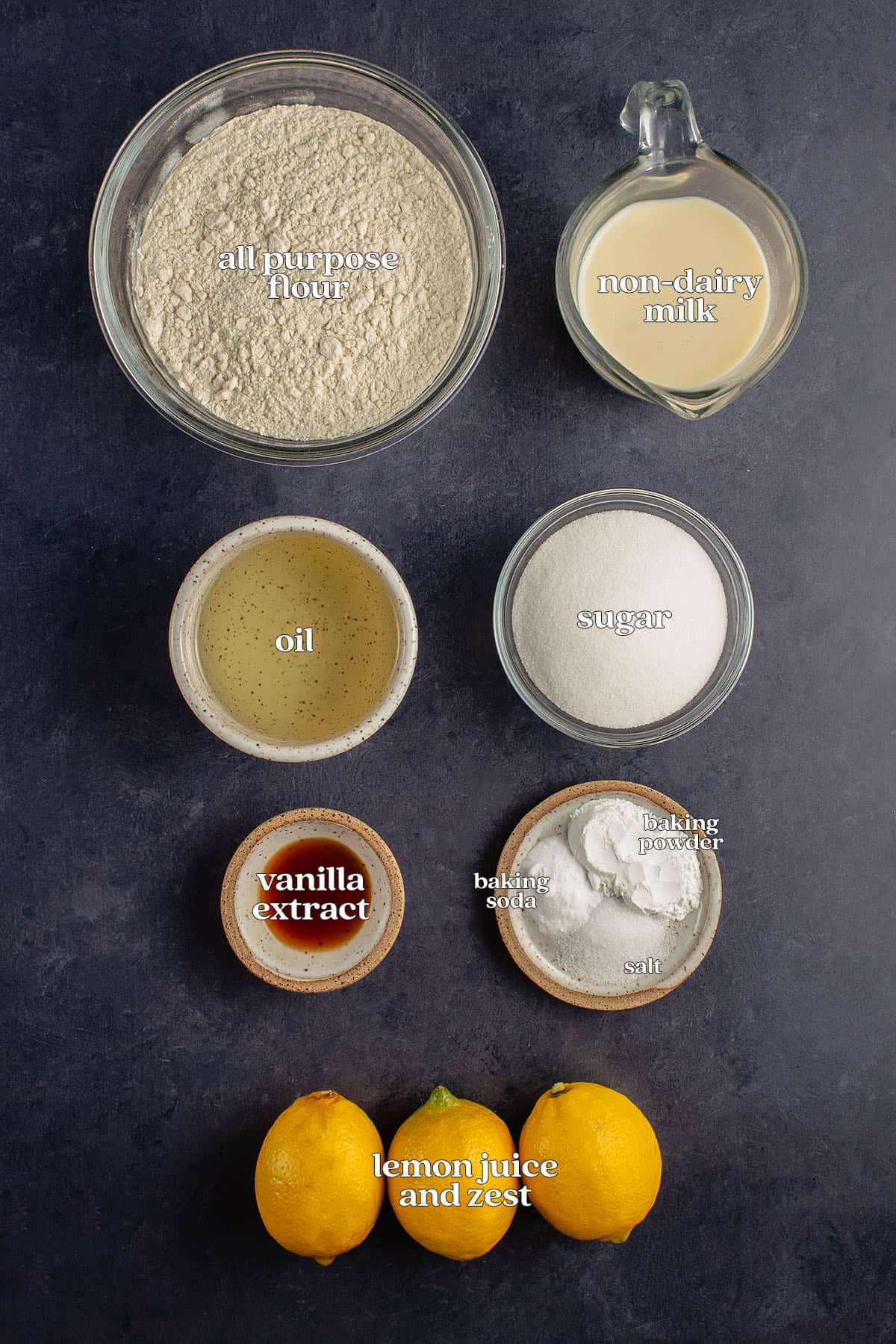 labeled ingredients for vegan glazed lemon loaf on blue backdrop: all purpose flour, fresh lemons, non-dairy milk, oil, sugar, vanilla extract, baking powder, baking soda, and salt