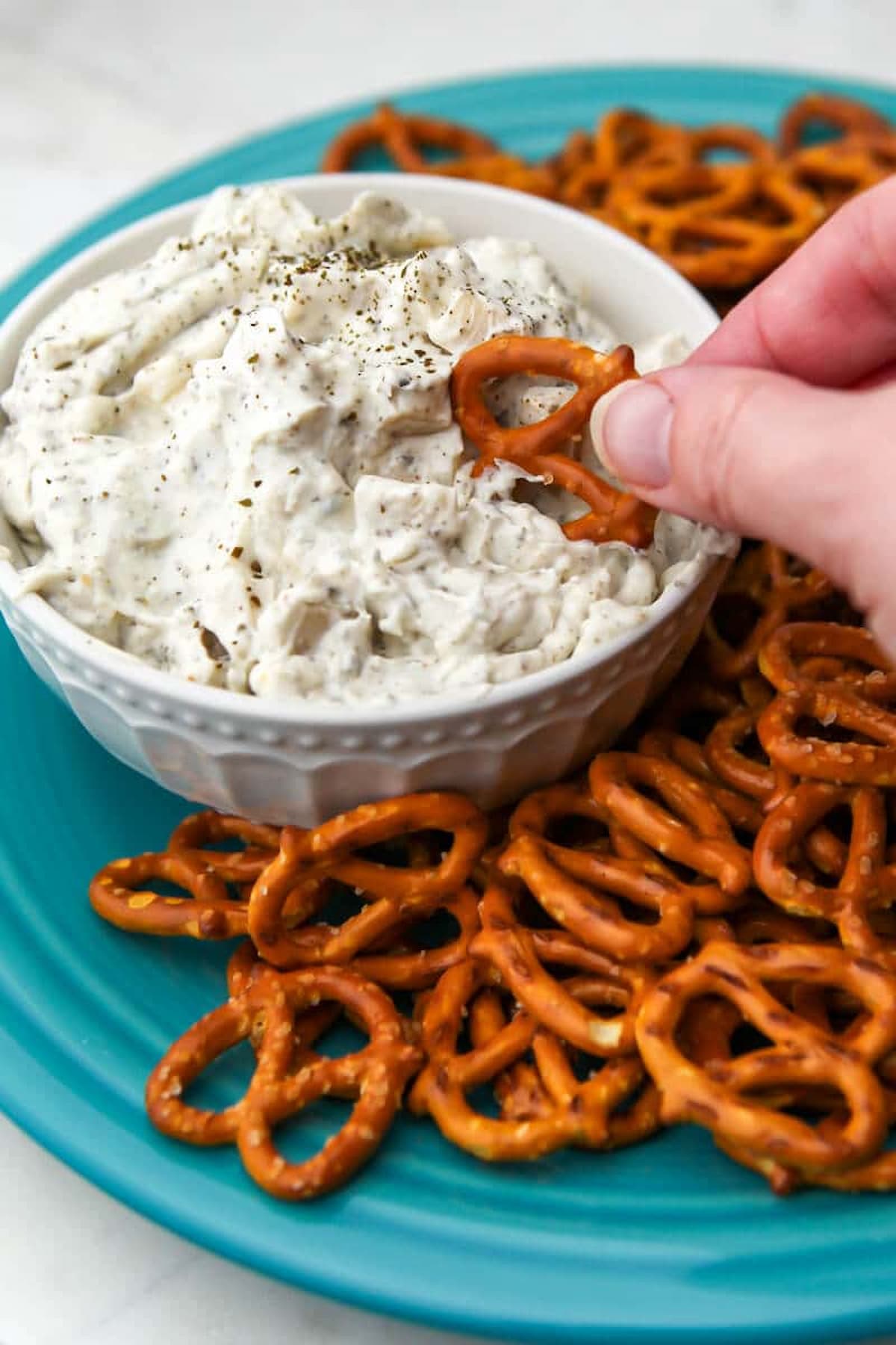 A hand dipping a pretzel into a bowl of vegan clam dip.