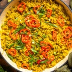 a white pot of garlic turmeric rice atop a table with red chilis and green onions garnished on the top.