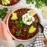 A bowl of vegan chili topped with fresh herbs, vegan sour cream, avocado slices, and a hand with a spoon about to scoop some up.