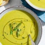 An overhead photo of two bowls of asparagus soup, one with a spoon. Next to these is a small plate of lemon wedges.