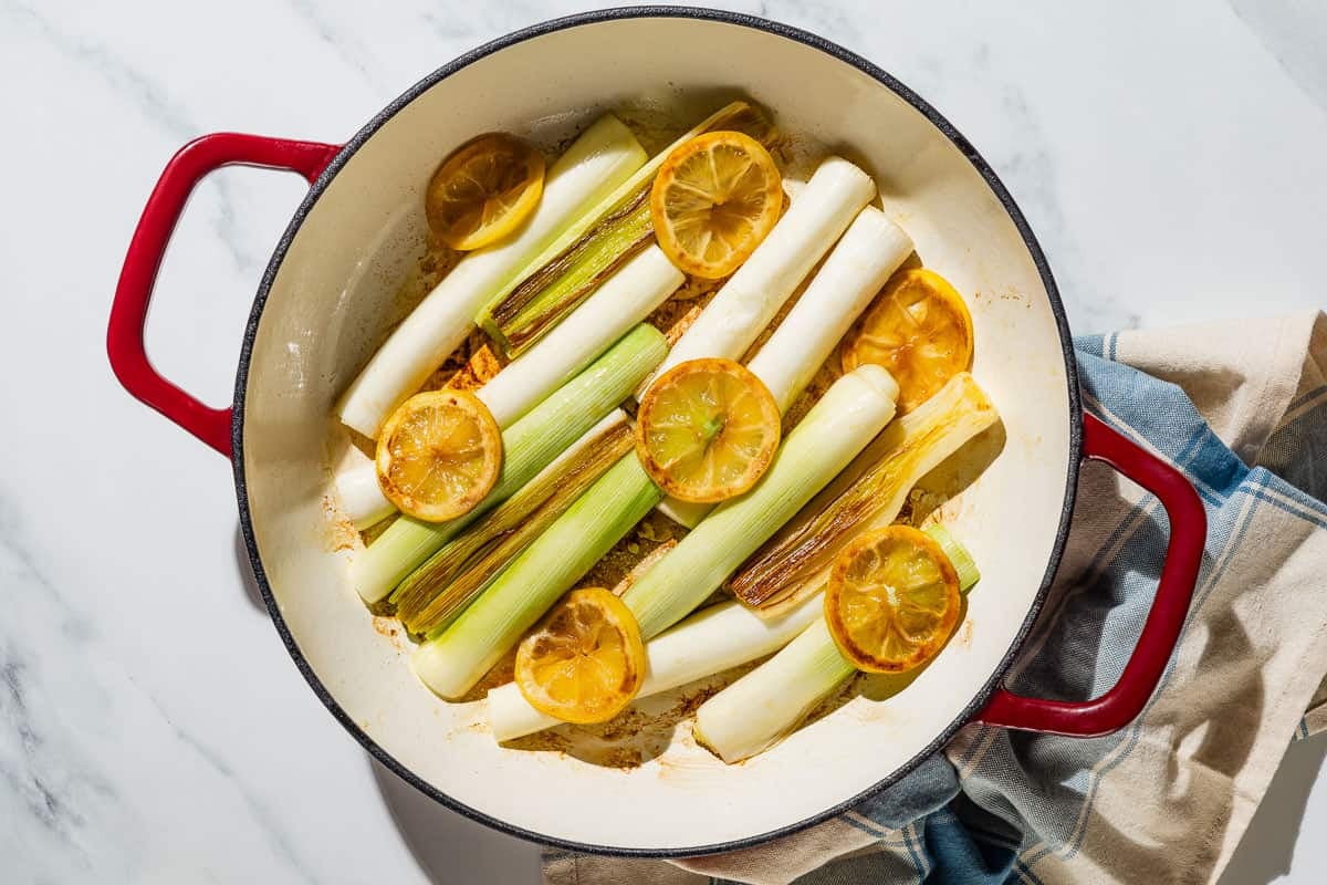 Leeks topped with lemon slices being braised in a skillet next to a cloth napkin.