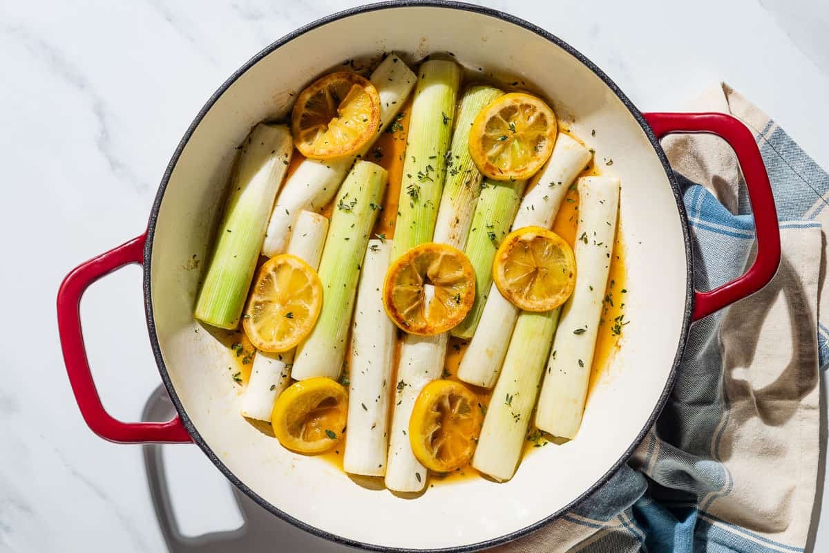 Leeks topped with lemon slices and herbs being braised in a skillet next to a cloth napkin.