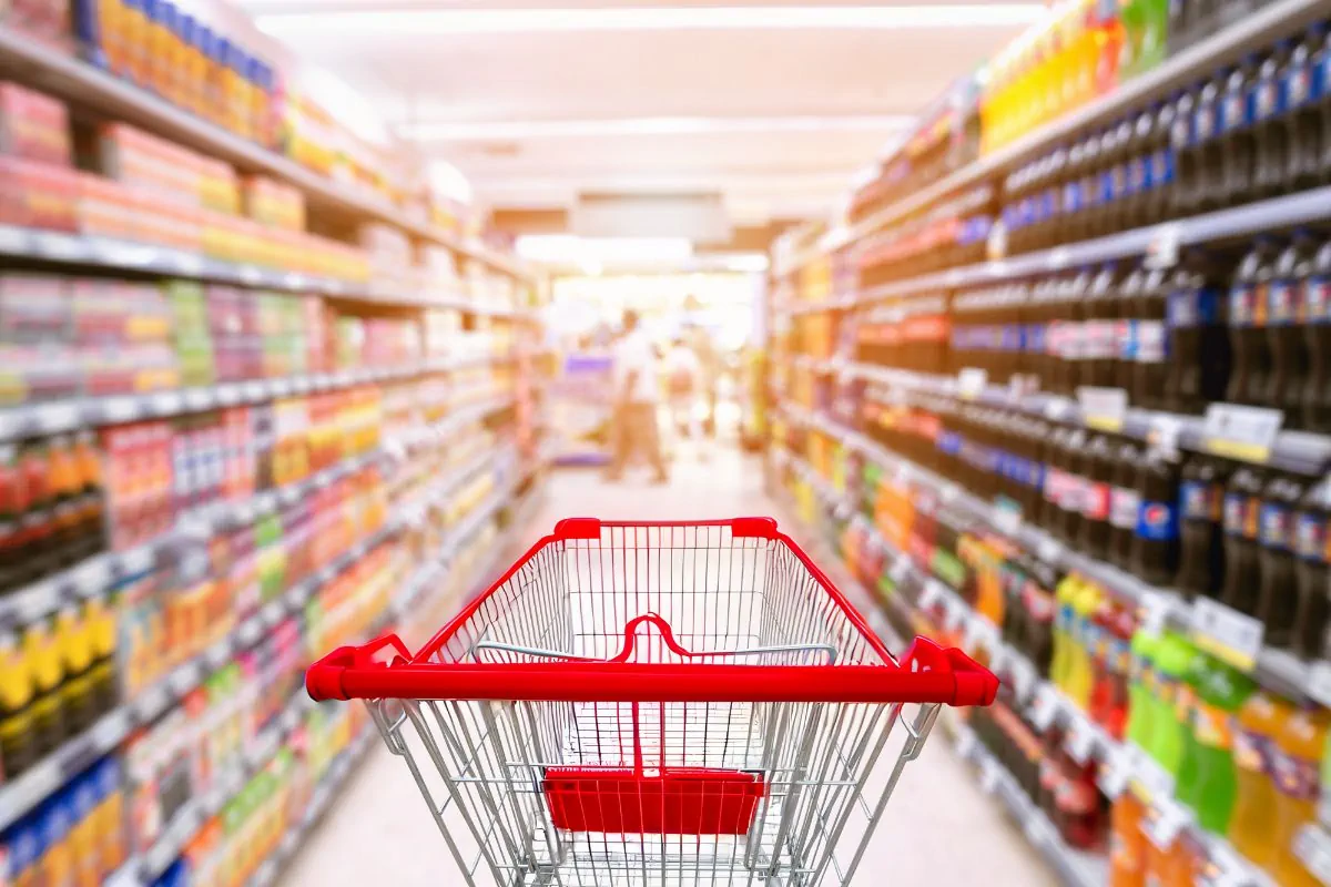 An empty shopping trolley between two shelves stocked with food items in a supermarket.